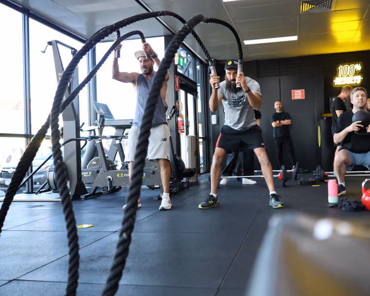 Two men exercising with battle ropes in a gym while others use weights and equipment in the background.