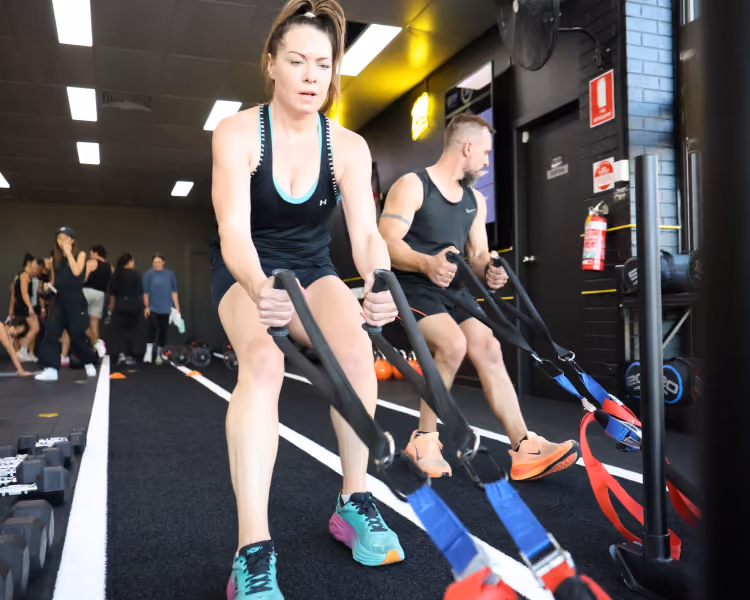 Two people using suspension trainers in a gym with others working out in the background.