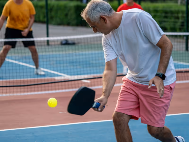 Man in a white shirt and pink shorts hitting a yellow pickleball with a black paddle on an outdoor court, with two other players in the background.