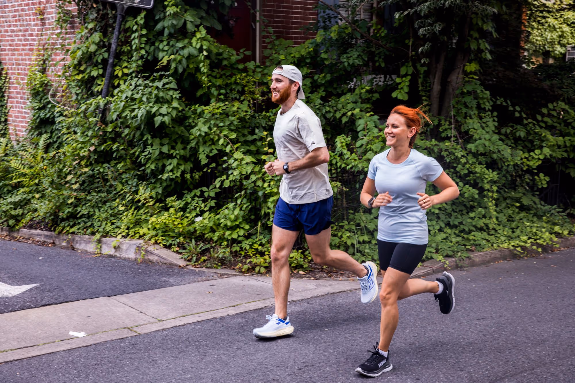 Man and woman jogging together on a street beside a wall covered with green ivy.