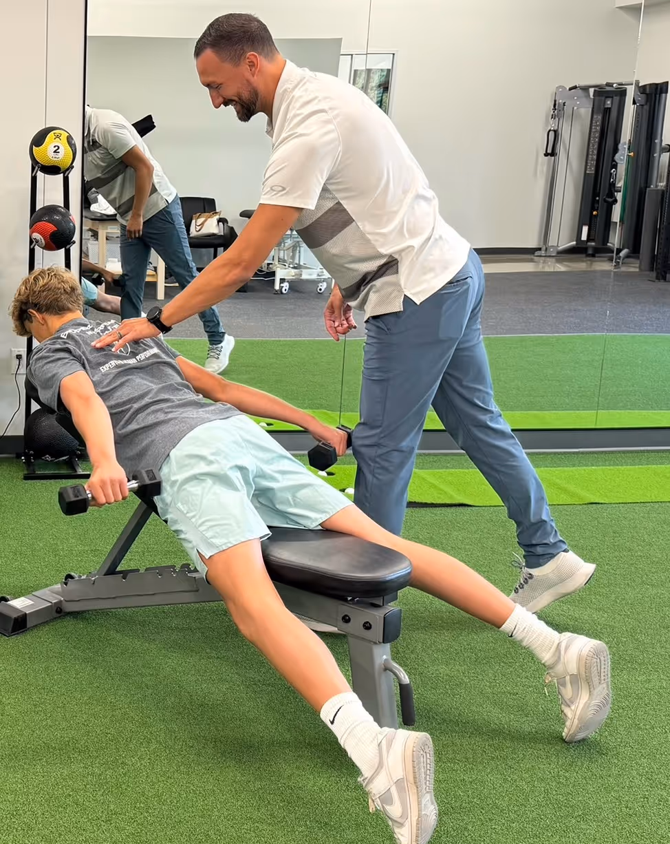 A trainer assists a young person doing dumbbell fly exercises while lying face down on an incline bench in a gym with green turf flooring.
