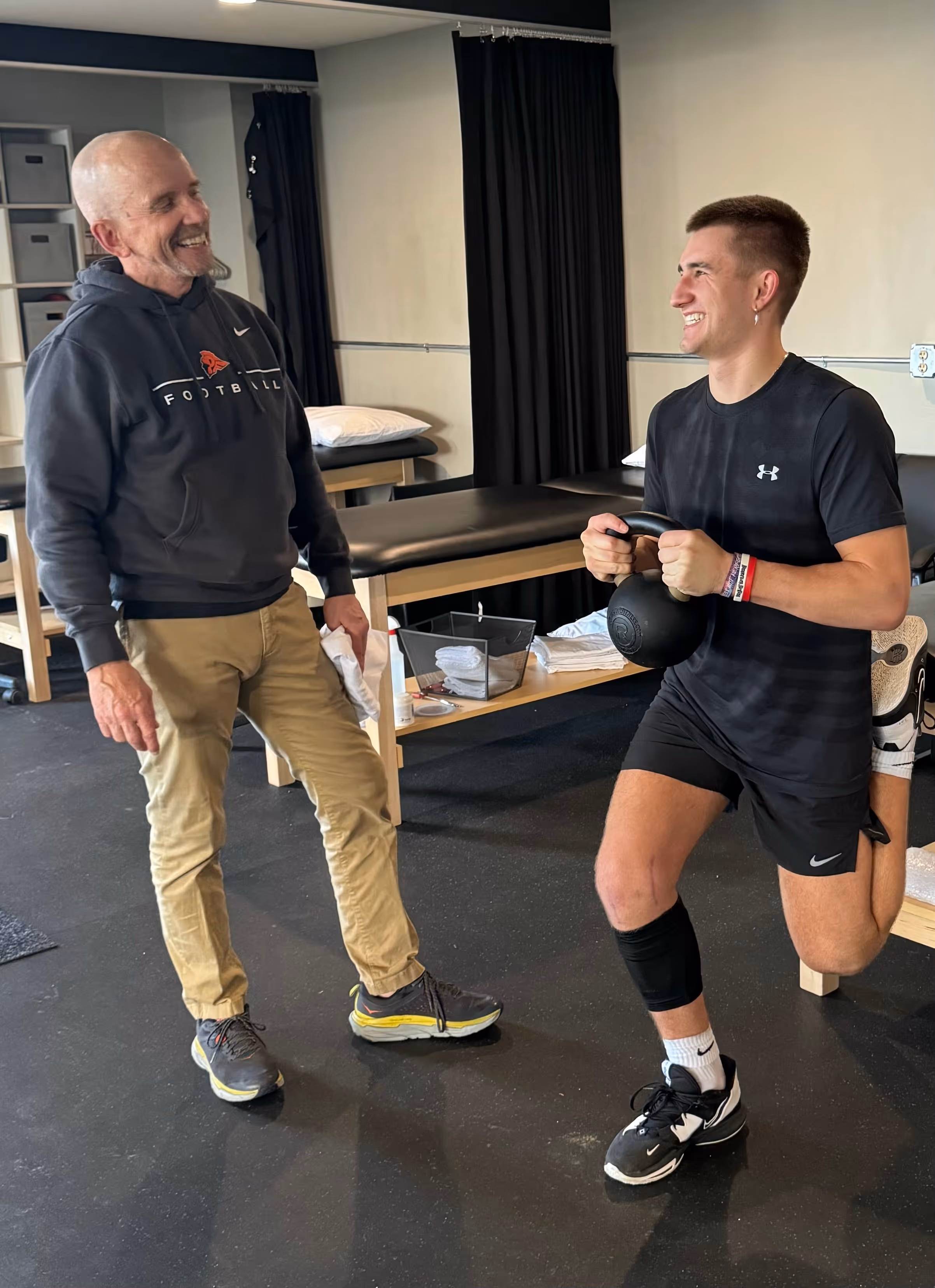 Young man holding a kettlebell and stretching one leg behind on a bench while an older man in casual clothes watches and smiles in a gym setting.