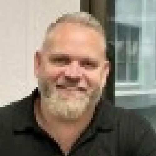 Blurred portrait of a man with light hair and a beard smiling, wearing a black shirt.