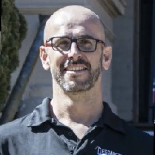 Smiling bald man with glasses and a trimmed beard wearing a black polo shirt outdoors.