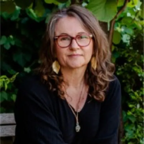 Woman with curly brown hair and glasses wearing a black top and leaf-shaped earrings, sitting outdoors with green foliage in the background.