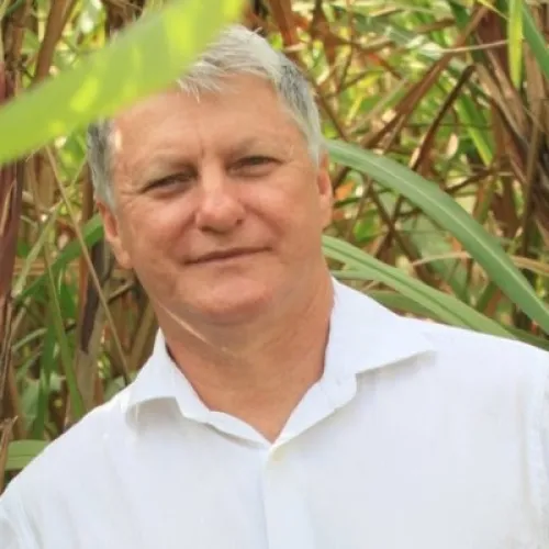 Middle-aged man with short gray hair wearing a white shirt, standing outdoors with green foliage in the background.