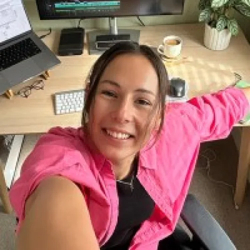 Smiling woman in a pink shirt taking a selfie while sitting at a desk with a laptop, keyboard, mouse, and a cup of coffee.