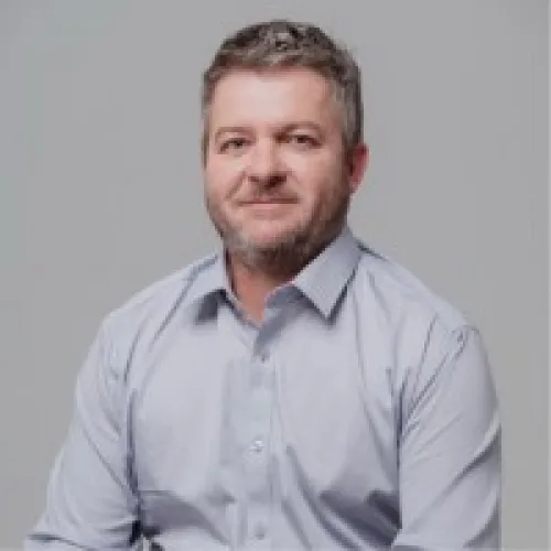 Portrait of a man with short hair and beard wearing a light blue button-up shirt against a gray background.