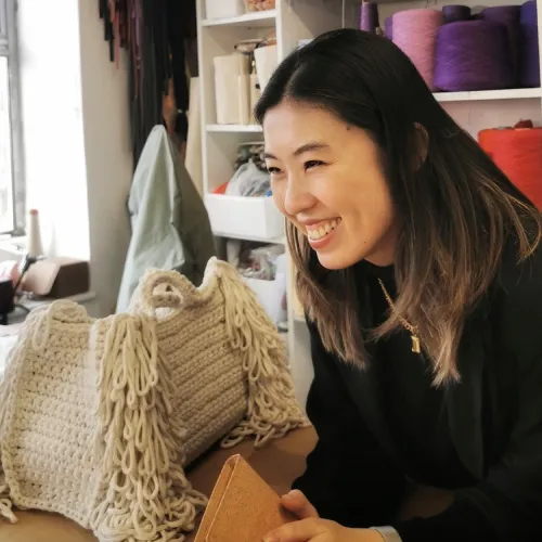 Smiling woman with shoulder-length hair leaning forward near a beige crocheted bag and colorful yarn spools on shelves.