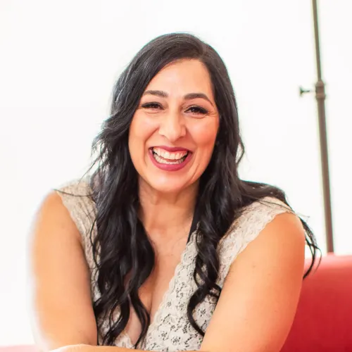 Smiling woman with long dark hair wearing a sleeveless beige lace top, sitting indoors against a light background.