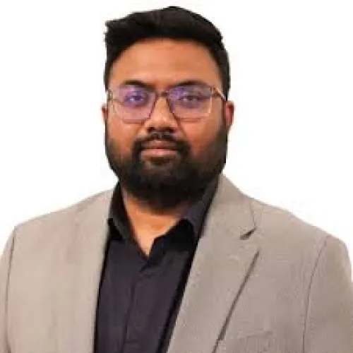 Man with glasses and beard wearing a beige blazer and black shirt against a white background.