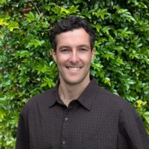 Smiling man with curly dark hair wearing a dark checkered shirt standing in front of green leafy bushes.