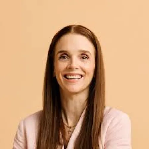 Smiling woman with long straight brown hair wearing a light pink blazer against a beige background.