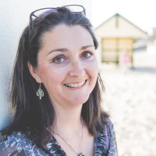 Smiling woman with dark hair, wearing glasses on her head and silver earrings, leaning against a light-colored wall outdoors.