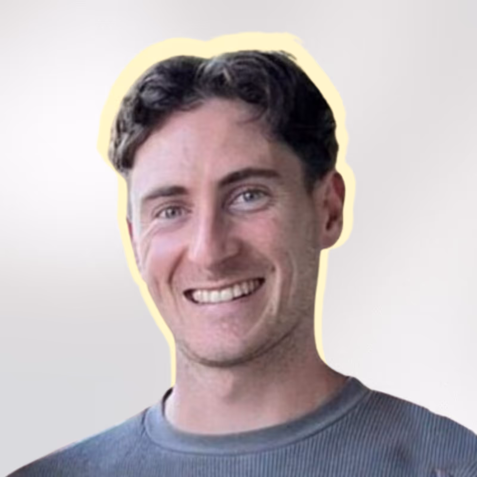 Smiling man with short wavy brown hair wearing a grey shirt against a light background with a soft yellow outline around his head.
