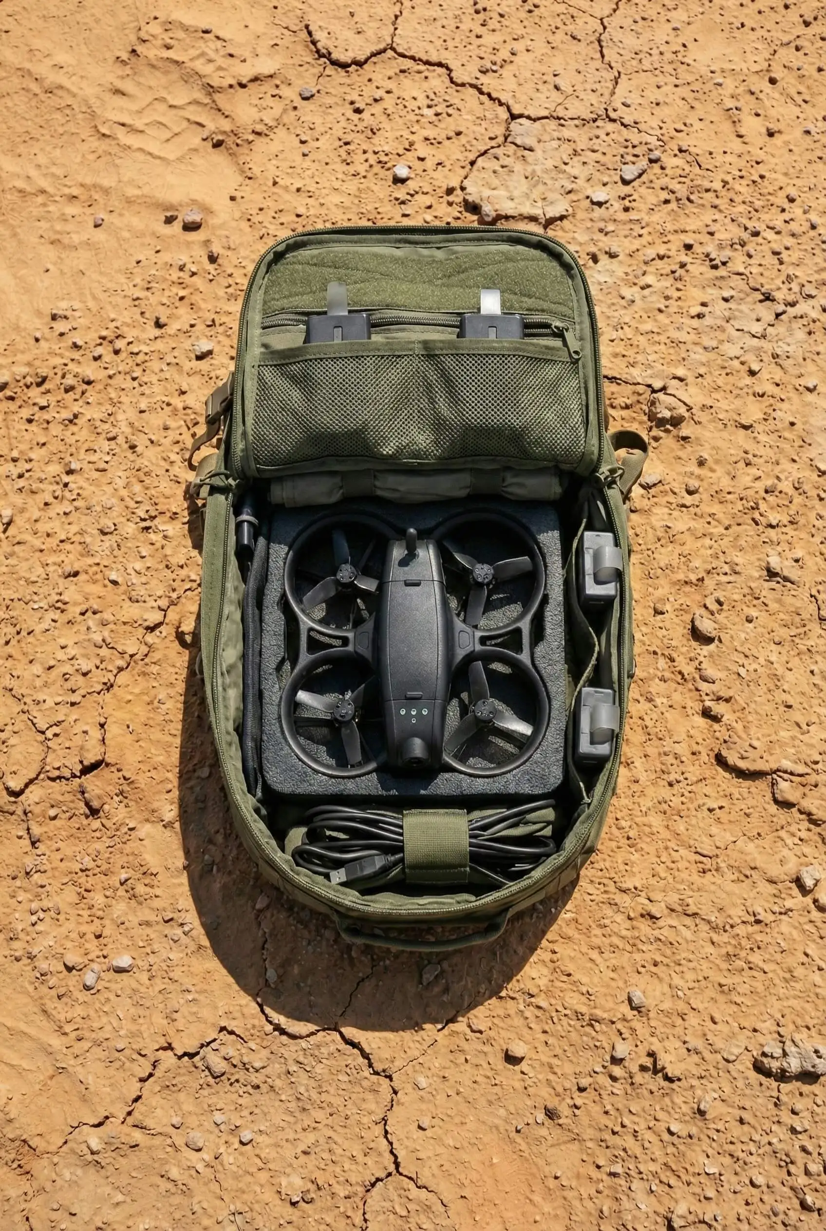 Open green backpack on dry cracked earth containing a compact drone, two batteries, and a USB cable.