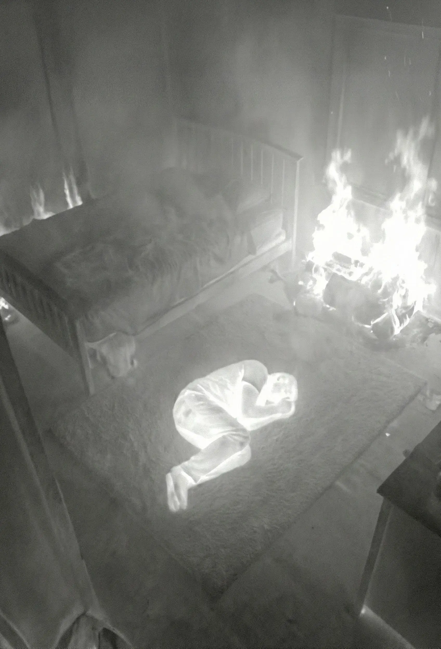 Thermal image showing a person curled up on a rug beside a bed, with a fire burning near the wall.