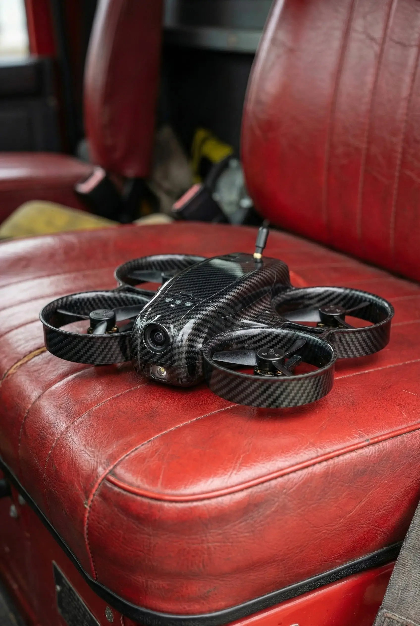 Small carbon fiber drone with four propellers resting on a red leather car seat.