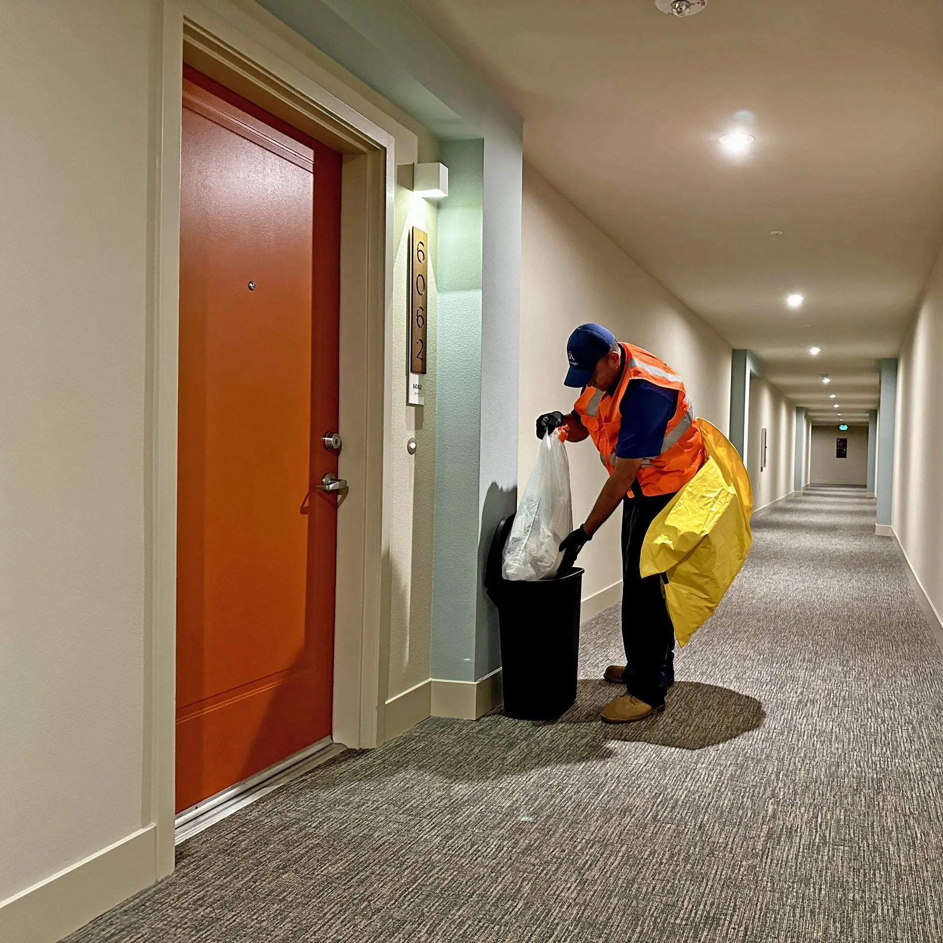 Maintenance worker in orange safety vest and blue cap emptying a trash bag into a black bin in a well-lit apartment hallway near an orange door numbered 6062.