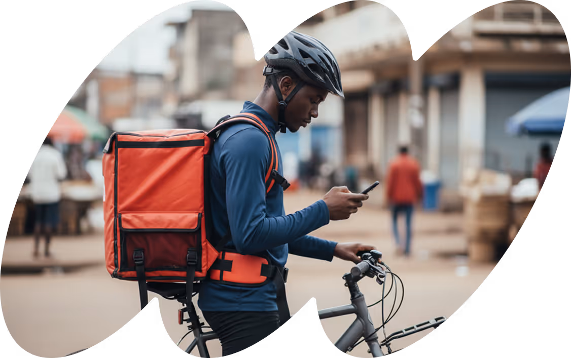 Delivery cyclist wearing a helmet and blue long-sleeve shirt checks a phone while holding a bicycle with a large red delivery bag on his back.