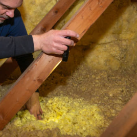 Person inspecting yellow insulation foam in an attic space under wooden beams using a flashlight.