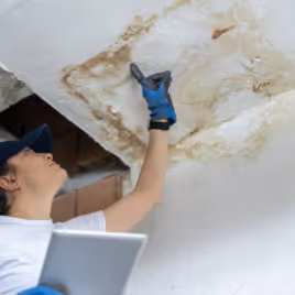 Person wearing blue gloves inspecting a large water stain on a ceiling while holding a tablet.