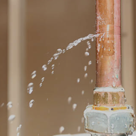 Close-up of a copper pipe leaking water from a joint with droplets spraying out.