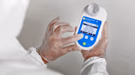 Person wearing protective gloves holding and examining a handheld electronic device with a barcode and screen display.