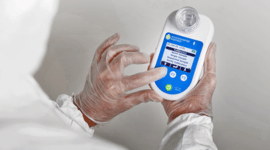 Person wearing protective gloves holding and examining a handheld electronic device with a barcode and screen display.