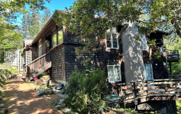 A wooden cabin surrounded by trees with a small wooden bridge and a dirt pathway in a forested area.