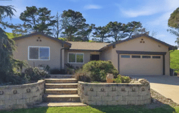 Single-story beige house with a two-car garage, landscaped front yard, stone retaining wall, and steps leading to the entrance.