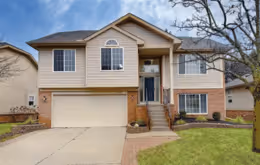 Two-story suburban house with beige siding, a two-car garage, small front porch, and landscaped yard with a tree.