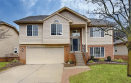 Two-story suburban house with beige siding, a two-car garage, small front porch, and landscaped yard with a tree.