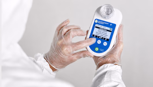 Person wearing protective gloves holding and examining a handheld electronic device with a barcode and screen display.