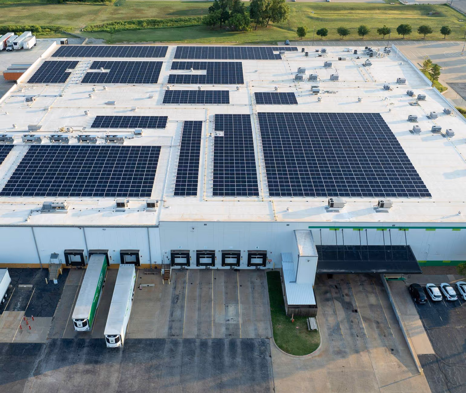 Aerial view of a large warehouse with multiple solar panel arrays on the flat white roof and trucks parked at loading docks.