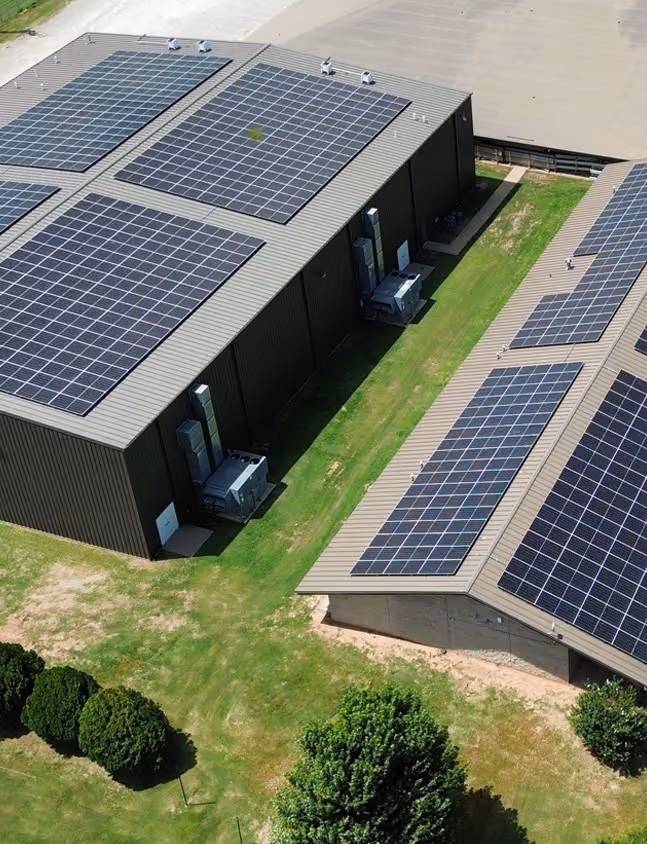 Aerial view of industrial buildings with solar panels installed on the roofs surrounded by green grass and trees.