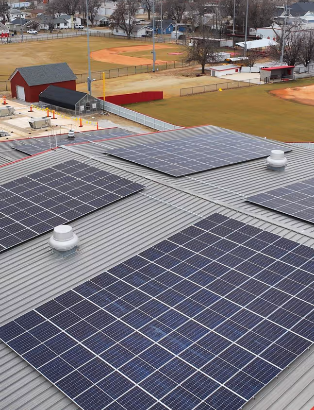 Solar panels installed on a corrugated metal roof overlooking a baseball field and a neighborhood with houses and trees.
