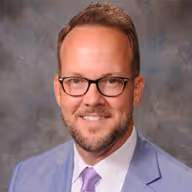 Smiling man with short hair, glasses, beard, wearing a light blue suit and purple tie against a gray background.