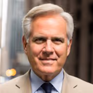 Portrait of a middle-aged man with gray hair wearing a beige suit jacket and blue shirt, smiling slightly.