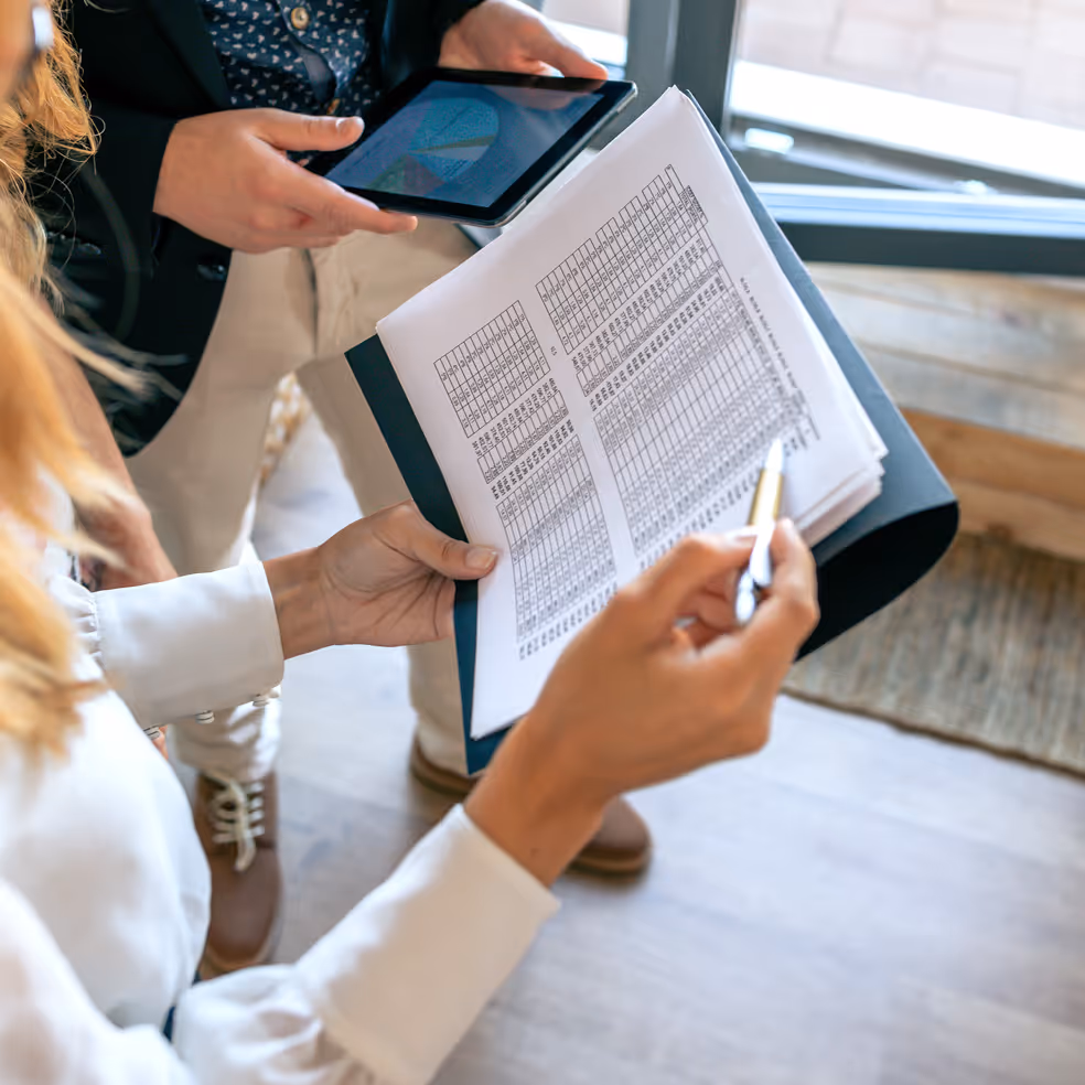 Two people reviewing printed spreadsheets and a tablet displaying a pie chart in a bright office.