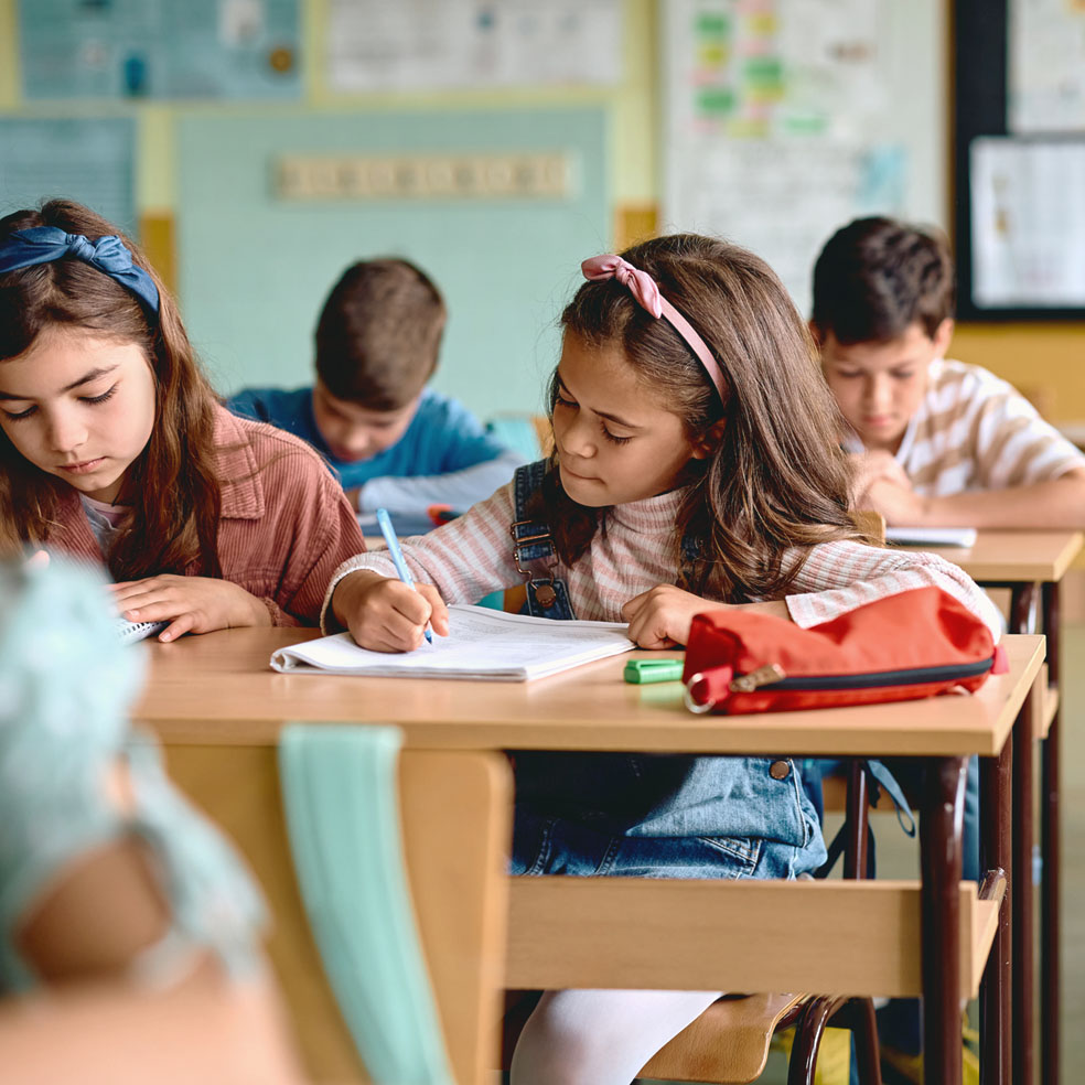 Four children sitting at classroom desks focused on writing in notebooks.