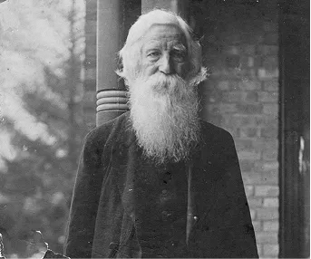 Black and white portrait of an elderly man with a long white beard standing outside near a column and brick wall.