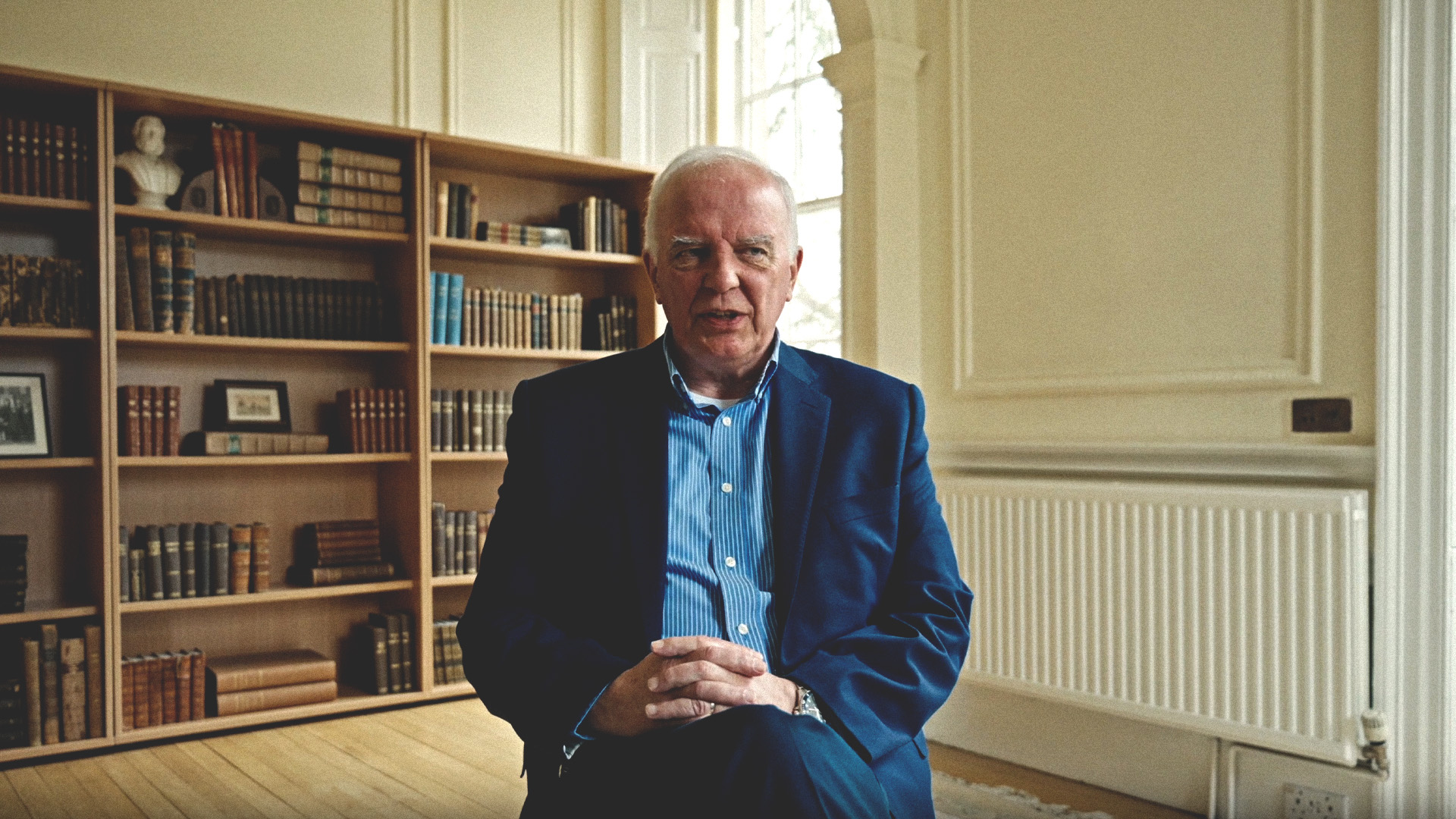 Ian Hamilton, in a blue suit, is sitting with clasped hands in a room with bookshelves and a window behind him.