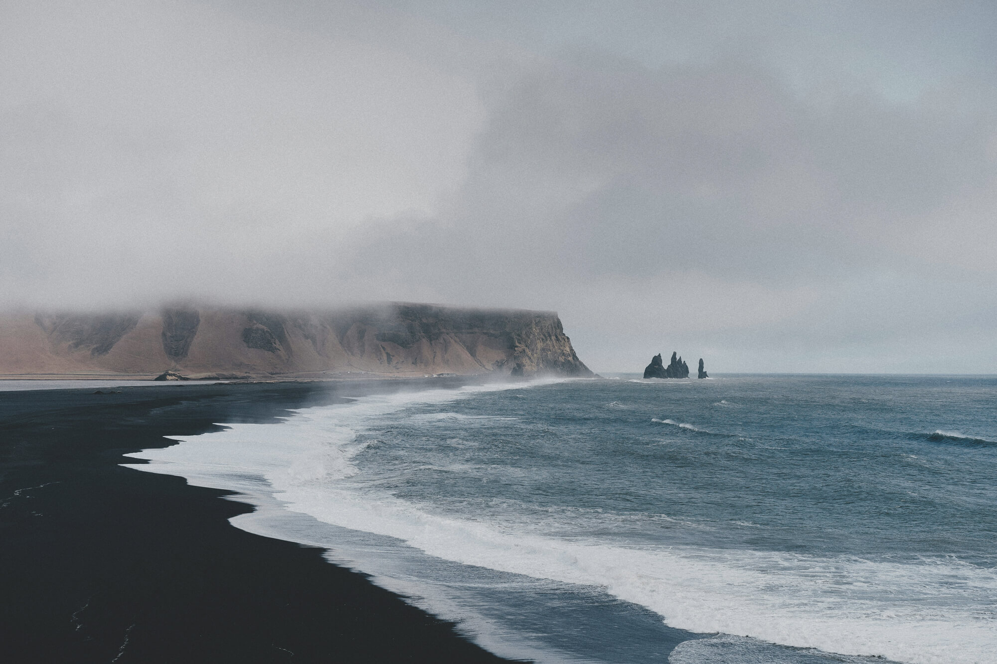 Black sand beach with waves crashing along the shoreline, cloudy sky, and rocky cliffs partially covered by fog in the distance.