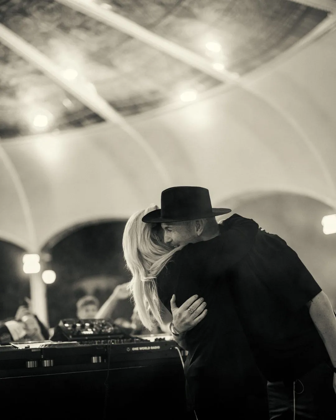 Black and white photo of a man wearing a hat and a woman with long hair hugging near DJ equipment under a tent.