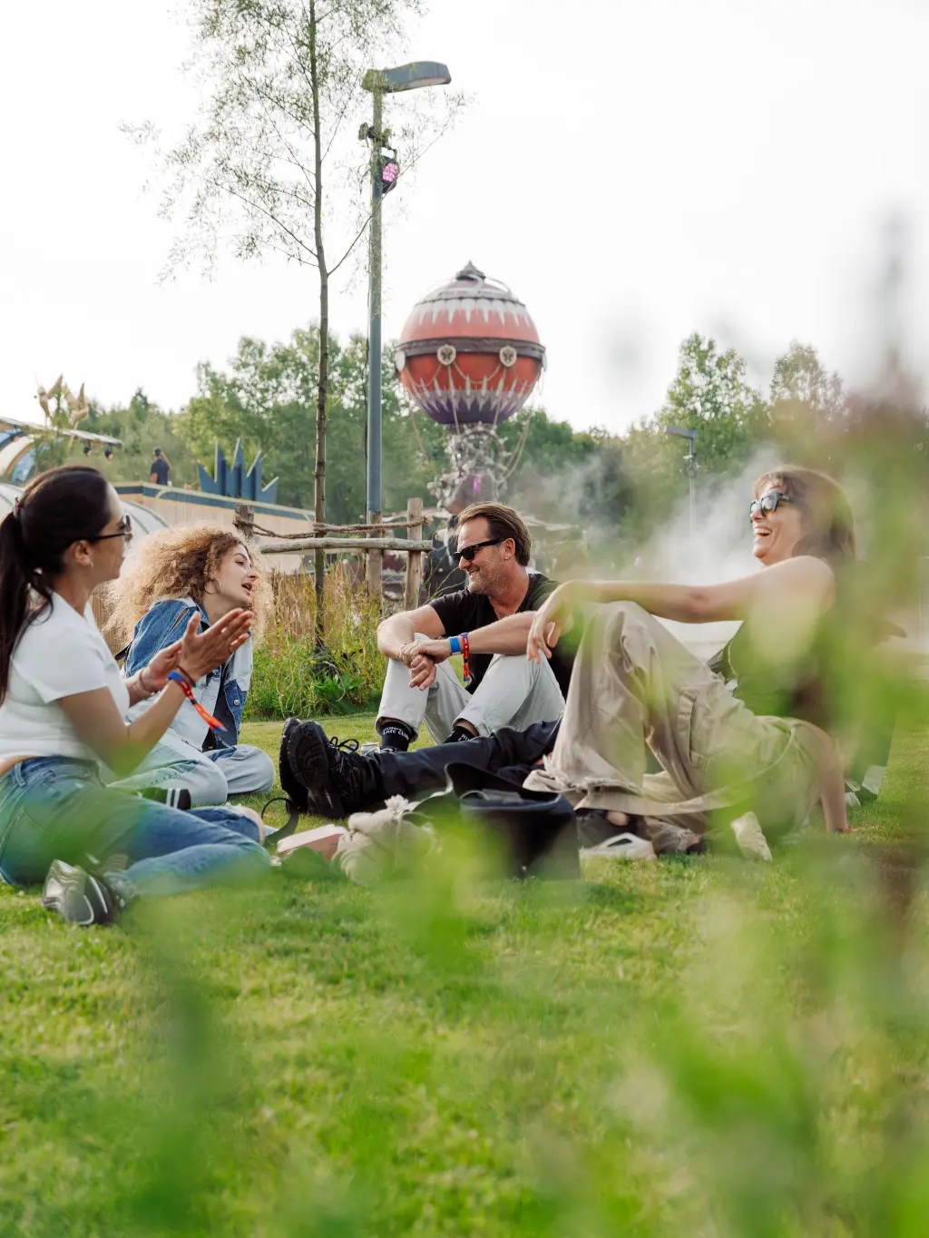 Four people sitting on grass in a park, chatting and laughing with trees and a decorative balloon structure in the background at the Love Tomorrow Summit