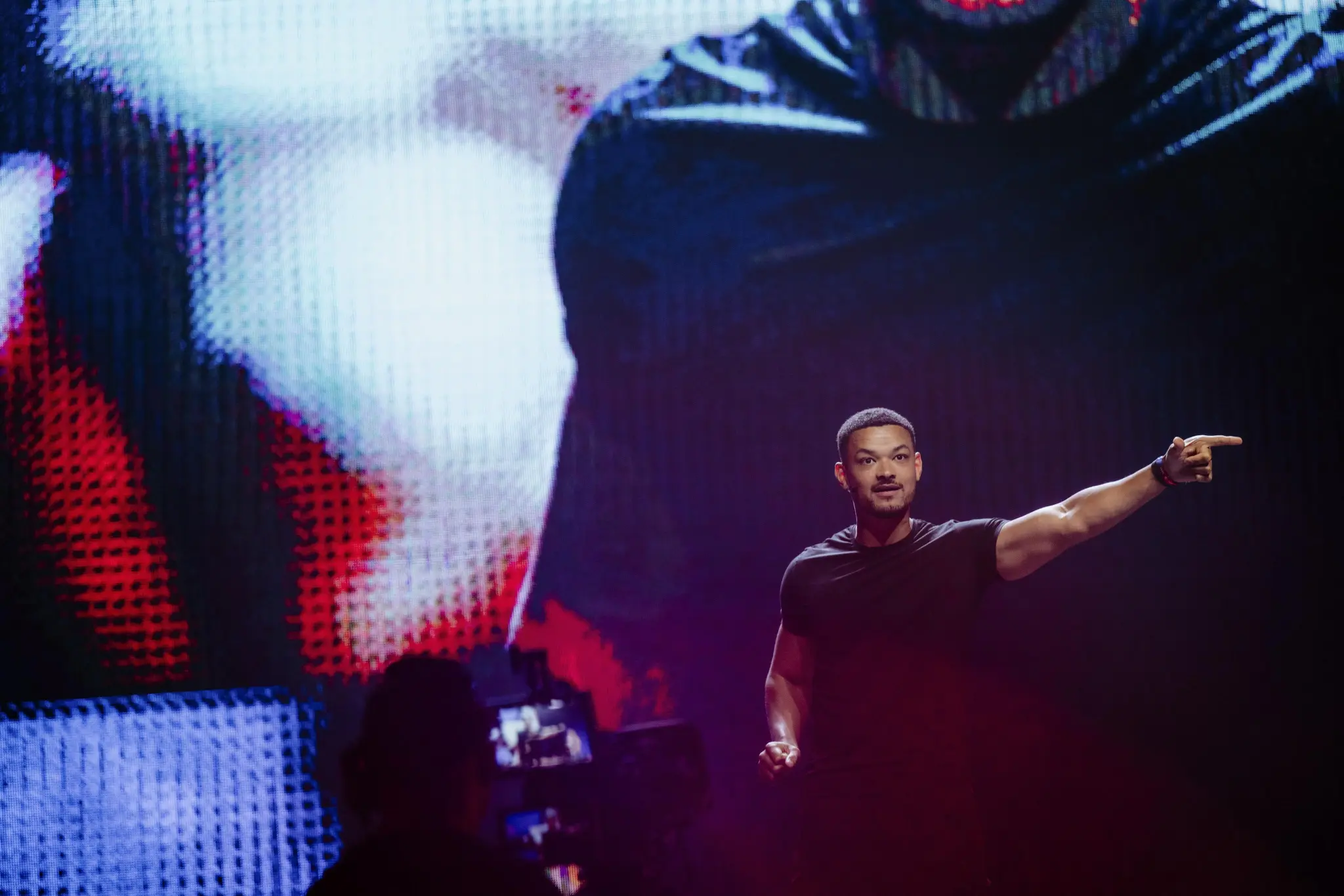 Steven Bartlett in black t-shirt on stage pointing to the right with a large colorful screen behind him at the Love Tomorrow Summit