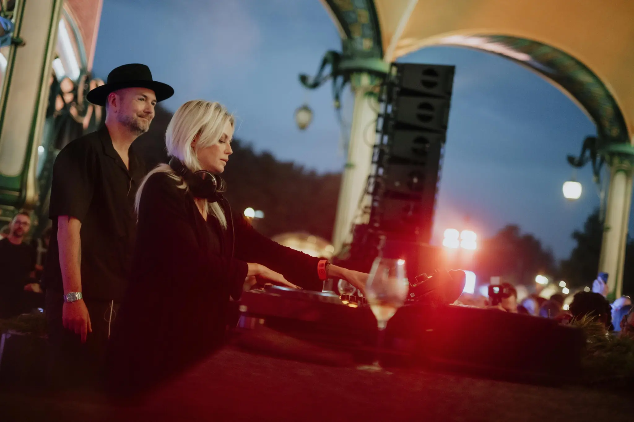 Female DJ with headphones on her neck mixing music at an outdoor event at dusk, with a man standing behind her and a crowd in the background at the Love Tomorrow Summit