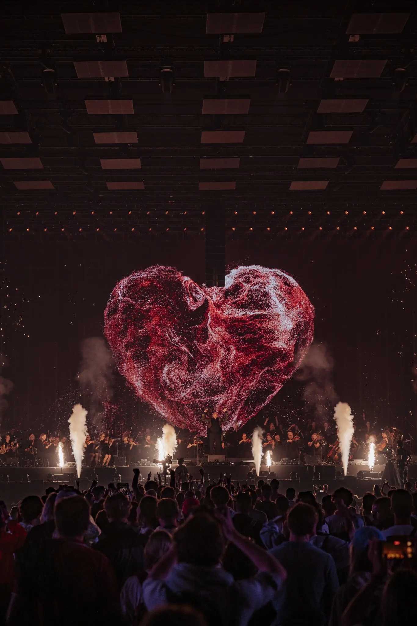 Crowd watching a live orchestral performance with a large red heart-shaped digital display and pyrotechnics on stage at the Love Tomorrow Summit