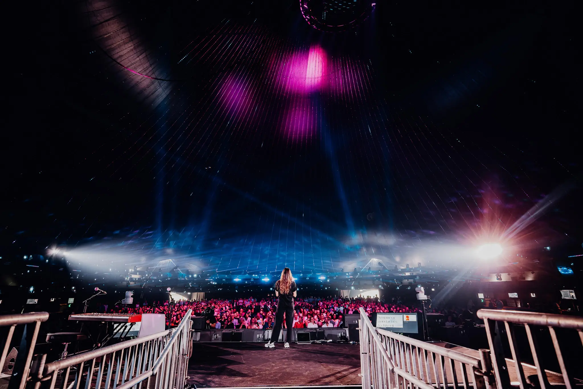 Person standing on stage facing a large audience under a dome ceiling with purple and blue lights at the Love Tomorrow Summit